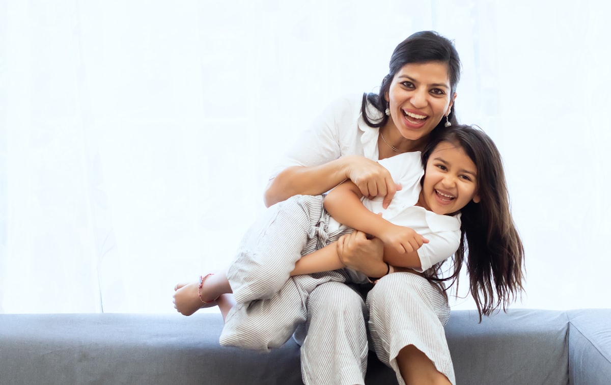 Stock photograph: a mother laughs while hugging her young daughter on a sofa beside a bright window.