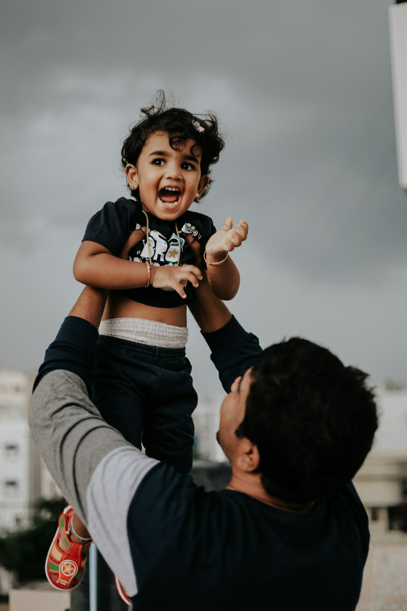 Stock photograph: a father lifts his laughing toddler overhead on an open rooftop, apartment buildings and a grey monsoon sky behind them.