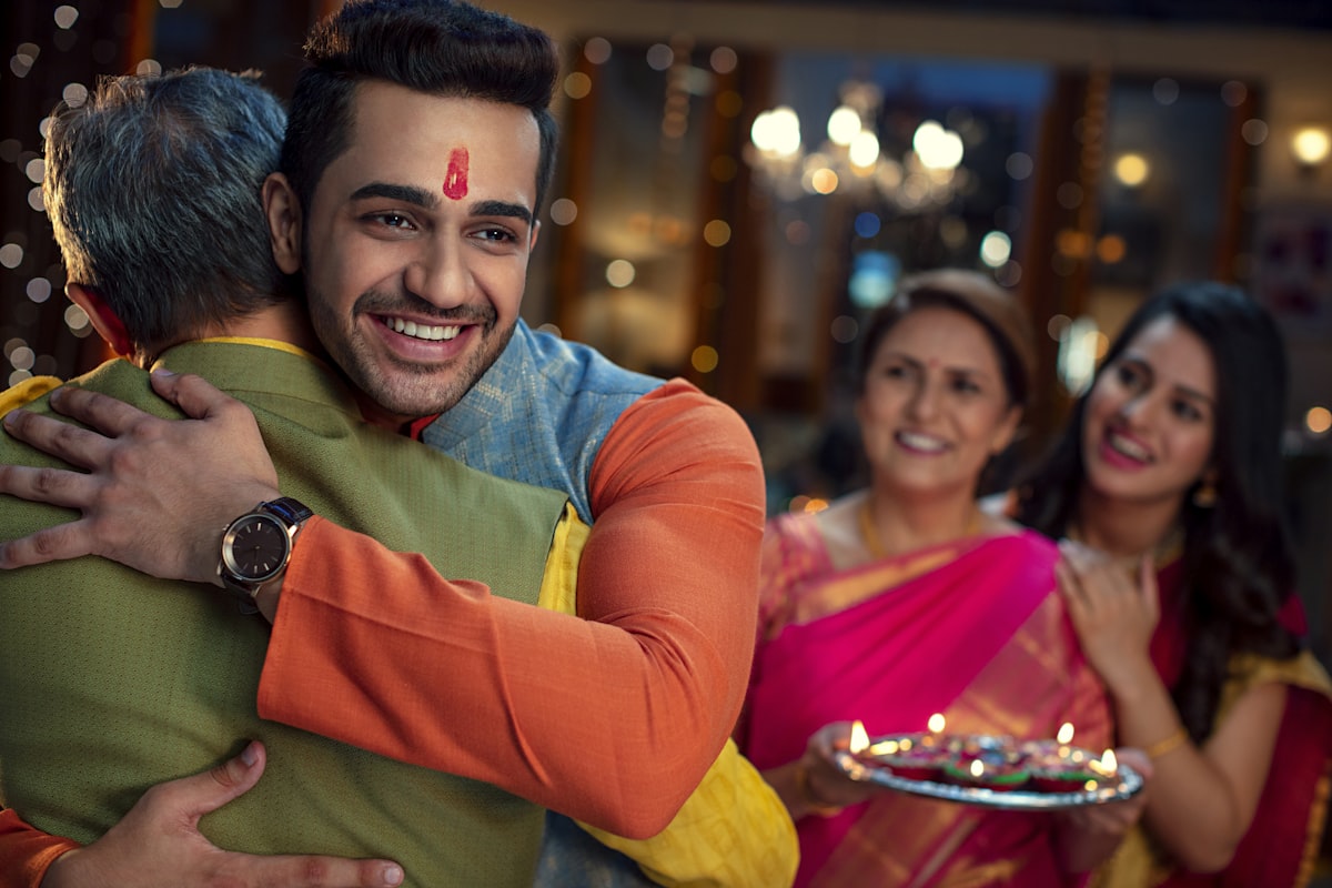 Stock photograph: a young man in a festive kurta embraces an older man at a family gathering, two women with a lamp-lit thali smiling behind them.