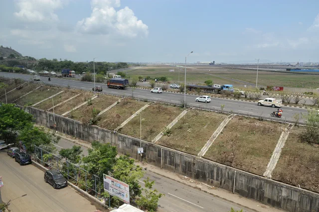 Trucks on the highway below the compound wall, the Navi Mumbai International Airport site beyond — Dhruva, Karanjade