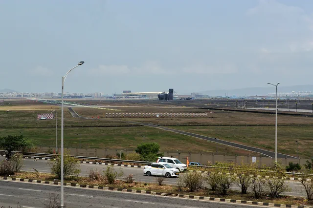 Airport terminal building and taxiway across the boundary road, past street lights and parked cars — Dhruva, Karanjade
