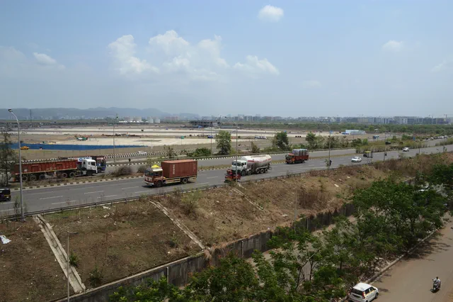 A line of red trucks on the highway, with the flat airfield and the hills behind them — Dhruva, Karanjade