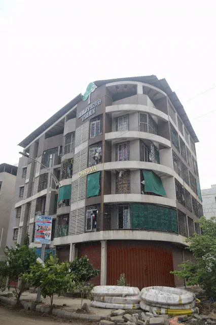 Curved corner elevation in grey and white, green balcony panels above shuttered shops — Dhruva, Karanjade
