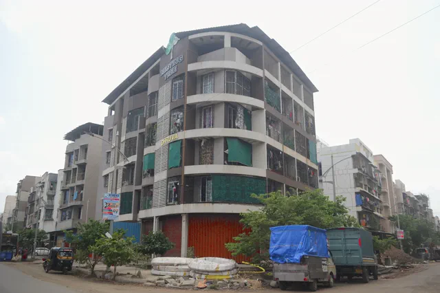 The corner plot seen along the street, past parked trucks and the neighbouring buildings — Dhruva, Karanjade