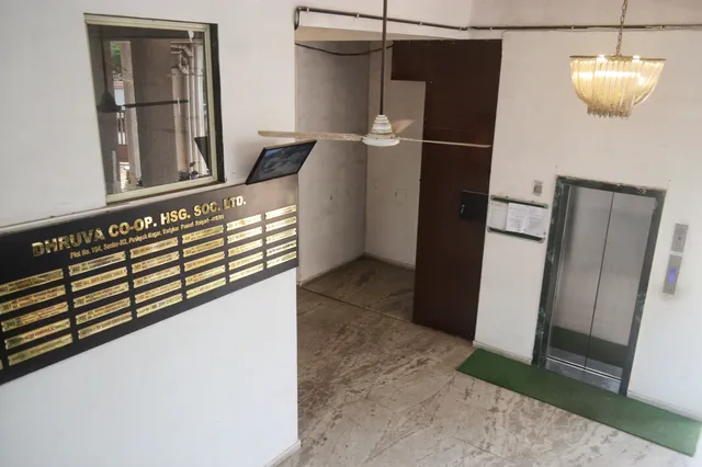 Brass society name board beside the lift landing in the entrance lobby, under a pendant light — Dhruva, Karanjade