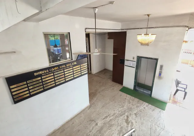 Lift doors, name board and window across the tiled entrance lobby, ceiling fan above — Dhruva, Karanjade