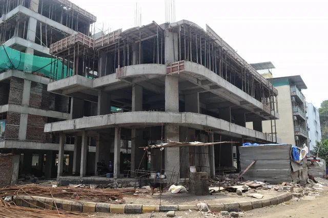 The concrete frame with green netting on the upper floors, rebar and materials stacked below — Prithvi, Karanjade