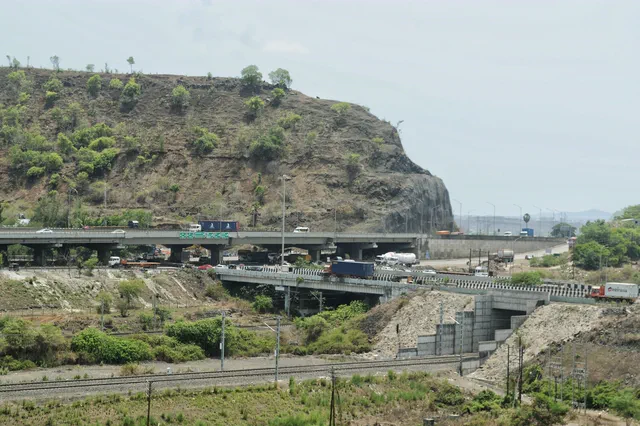 The flyover curving under the quarried cliff face, retaining walls and the rail line beneath it — Rudra, Karanjade