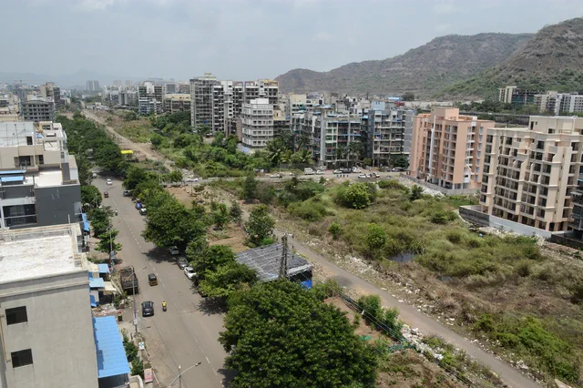 A broad road lined with apartment blocks, open plots to either side and the hills behind — Rudra, Karanjade