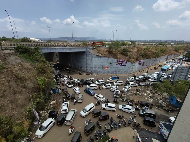Cars and motorcycles packed onto the open ground beside the underpass retaining wall — Rudra, Karanjade