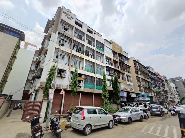 The building along the street with its neighbours, shops at pavement level and cars parked nose-in — Rudra, Karanjade