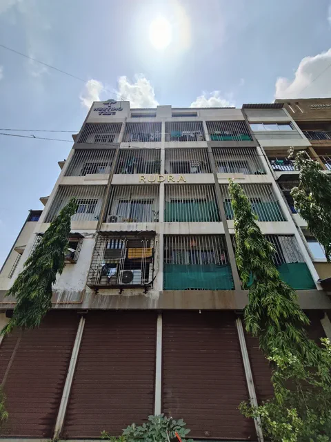 Looking up the elevation into the sun, climbers on the balconies and the shutters closed below — Rudra, Karanjade