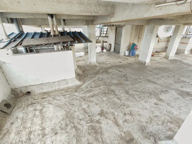 Bare concrete floor and columns on the covered rooftop level, roofing sheets stacked by the parapet — Rudra, Karanjade