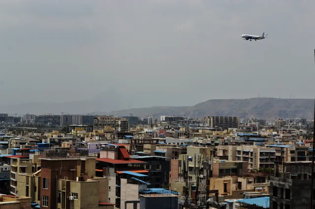 An airliner descending over the packed rooftops and water tanks of the town — Shaurya, Karanjade