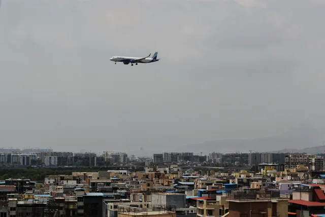 An airliner high over a hazy skyline of rooftops, tower cranes standing among the far blocks — Shaurya, Karanjade