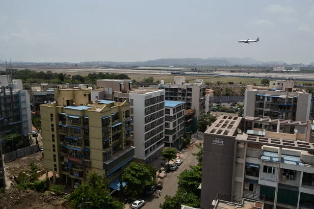 Mid-rise blocks along the street below, the airfield beyond them and an aircraft passing above — Shaurya, Karanjade