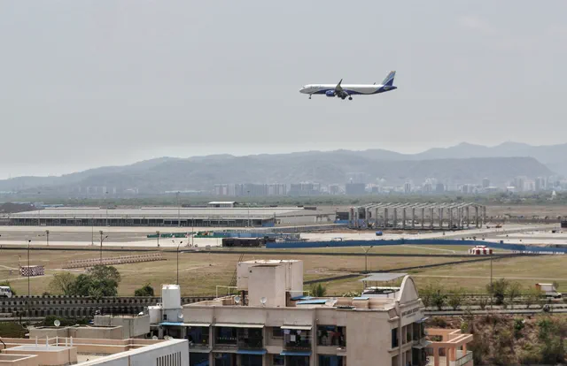 An airliner over the terminal pier and its jet bridges, water tanks on the rooftops below — Shaurya, Karanjade