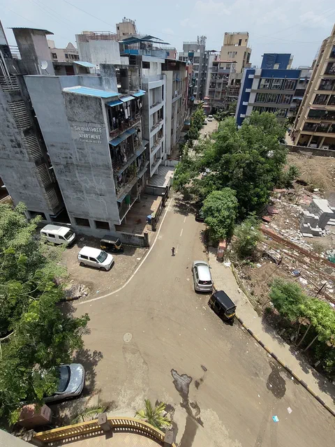 Straight down into the street between the blocks, cars parked along it and trees in the gaps — Shaurya, Karanjade