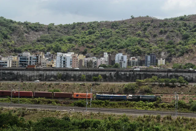A freight train on the line below the wooded hillside, apartment blocks along its foot — Shikhar, Karanjade