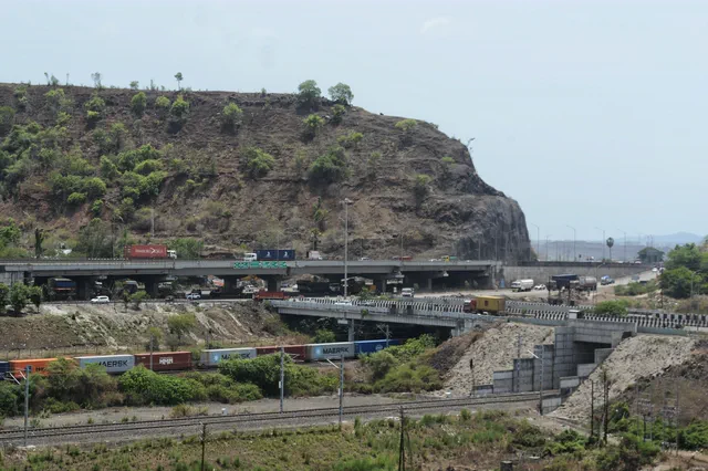 Traffic crossing the flyover beneath the quarried cliff face, container wagons on the line below — Shikhar, Karanjade