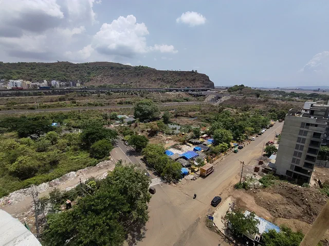 An unmade road curving past open ground, a tower under construction at its edge and hills beyond — Shikhar, Karanjade