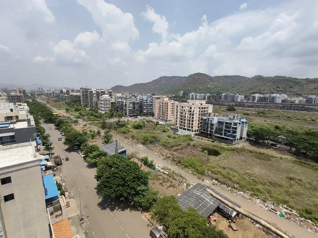 A tree-lined road running out between apartment blocks toward the hills, open ground either side — Shikhar, Karanjade