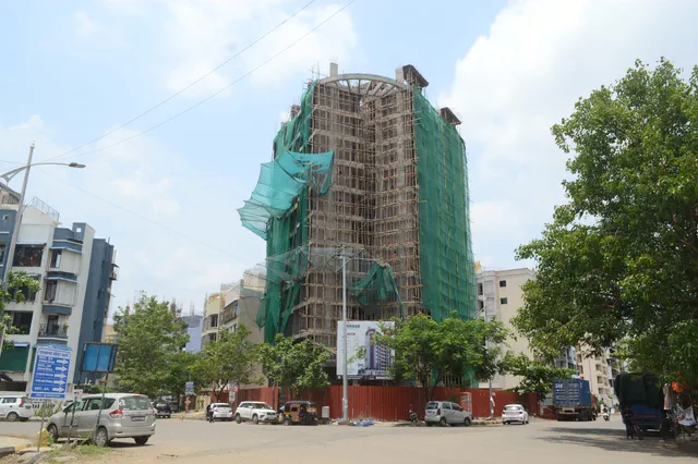 Seen from the street corner past the trees: scaffolding, green netting and the site hoarding — Shikhar, Karanjade