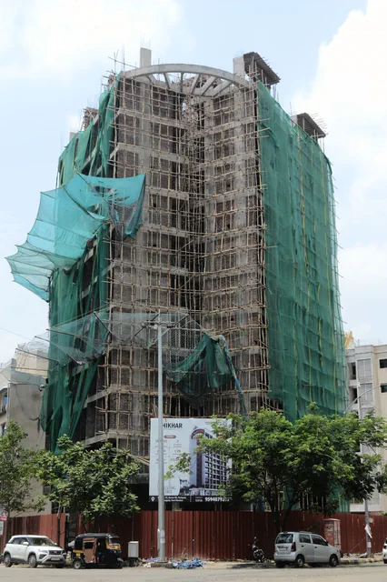 The full height of the building under scaffolding, netting billowing from the upper floors — Shikhar, Karanjade